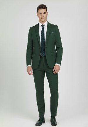 Hombre de pie con un traje verde oscuro, camisa blanca, corbata oscura con estampado y zapatos de vestir verdes a juego, sobre un fondo claro y sencillo.