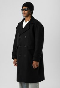 Zwarte double-breasted jas gemaakt van een dikke stof, met oversized revers, grote knopen en opgerolde manchetten, gedragen over een wit overhemd.