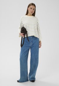 Pull ample à rayures crème et bleu clair, associé à un jean denim bleu à jambes larges. Tient un petit sac marron foncé à texture lisse.