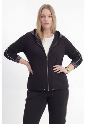 Vrouw met steil blond haar draagt een zwarte hoodie met ritssluiting en bijpassende broek met witte strepen, staand met haar handen in haar zij tegen een witte achtergrond.