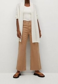 Kevyt, tekstuureiltaan valkoinen neuletakki istuvan valkoisen tanktopin päällä, yhdistettynä korkea vyötäröisiin, leveälahkeisiin beigen värisiin housuihin ja ruskeisiin flip-flop-sandaaleihin.