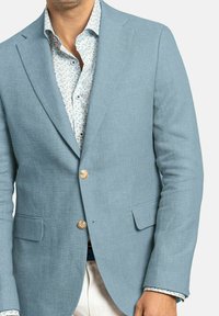 Blazer de lino azul claro con un acabado texturizado, dos bolsillos frontales y botones marrones. Lleva puesto sobre una camisa blanca con un patrón y pequeños acentos florales.