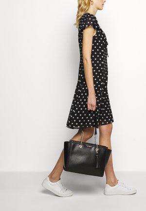 Borsa a tracolla in pelle nera con catena argentata e nappina, struttura rigida e texture liscia. Abbinata a un vestito a pois e sneakers bianche.