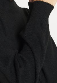 Un pull col roulé noir en tricot avec des manches longues, porté par une personne se touchant le visage d'une main.