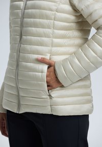 Lys beige puffy jakke med quiltet tekstur, slank pasform, lynlås foran og sidelommer. Materialet ser glat og skinnende ud.
