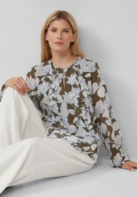 Bluse mit Blumenmuster und Rüschendetail in Olivgrün und Hellblau, mit langen Ärmeln und einem Ausschnitt mit Schlüsselhole. Kombiniert mit weißen Hosen.