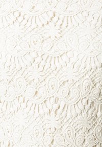 Spets tyg i off-white som har intrikata blommönster och svängda linjer, med en texturerad yta som ramar in öppna områden mellan designen.