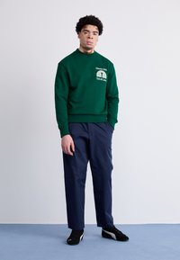 Sudadera verde con cuello redondo, con texto y logo en blanco. Combinada con pantalones azul marino y zapatillas negras. Diseño y ajuste simples.