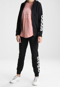 Černá mikina s kapucí na zip s bílým logem "adidas" na rukávu, v kombinaci se světle růžovým tričkem a černými teplákovými kalhotami se stejným logem. Bílé tenisky.