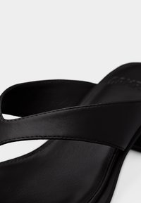 Zbliżenie na czarną skórzaną sandałkę z dwoma nakładającymi się paskami i widocznym przeszyciem na białym tle.