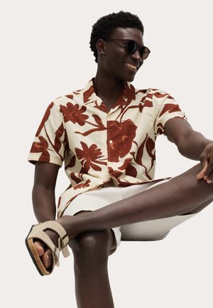 Ein Mann trägt eine Sonnenbrille, ein cremefarbenes Hemd mit großem braunem Blumenmuster, beige Shorts und beige Sandalen, sitzt mit übereinandergeschlagenem Bein.