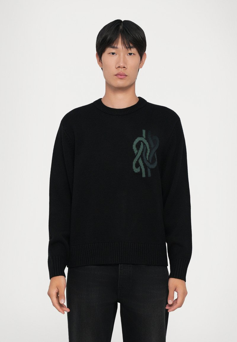 Young man with short black hair wearing a black sweater with a green abstract knot design on the chest and dark pants.