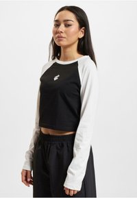 Crop top features black body and white long sleeves, made from soft fabric. Includes a small white logo on the chest.