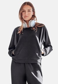 Sudadera negra de manga larga con un corte relajado, bolsillo frontal y material suave. Se lleva con grandes auriculares de diadema de un gris claro.