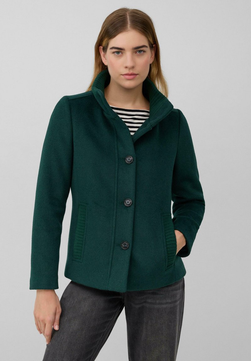 Dunkelgrüner Wollmischjacke mit fünf Knöpfen, übergroßem Kragen und gerippten Seitentaschen. Trägt man über einem schwarz-weißen gestreiften Oberteil.