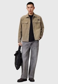 Beige cordjacka med två bröstfickor, ihop med en svart skjorta och slitna gråvida jeans. Håller en svart väska.