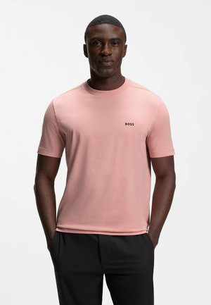 Uomo in piedi con le mani in tasca che indossa una t-shirt BOSS rosa chiaro e pantaloni neri su uno sfondo bianco uniforme.