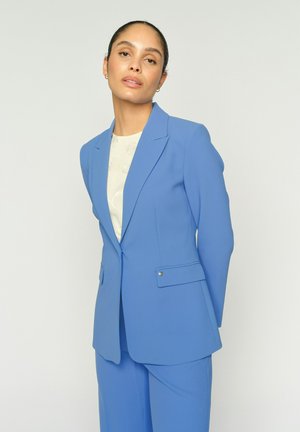 Vrouw met achterovergekamd haar, gekleed in een lichtblauw, op maat gemaakt pak en een witte top met patroon, staand tegen een effen achtergrond.