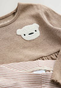 Body neonato beige a coste con una faccia di orso bianca e testurizzata sul petto, sovrapposto a pantaloni a righe beige e bianchi.