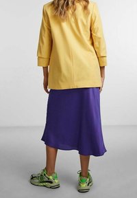 Żółta marynarka z rękawami do łokci na tle zwiewnej fioletowej spódnicy; zestawiona z szarymi i zielonymi butami sportowymi.