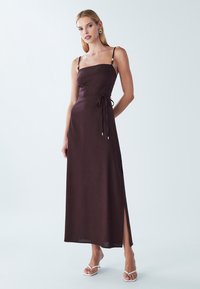 Abito lungo marrone scuro senza maniche con spacco laterale e cintura annodata in vita, con sottili spalline decorate con perline. Tessuto liscio e design minimalista.