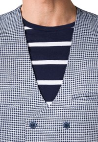 Dwurzędowa kamizelka w wzór houndstooth w odcieniach granatu i bieli, dopasowany krój, z niebieskimi guzikami i kieszenią na piersi. Pod spód założona jest koszula w paski.