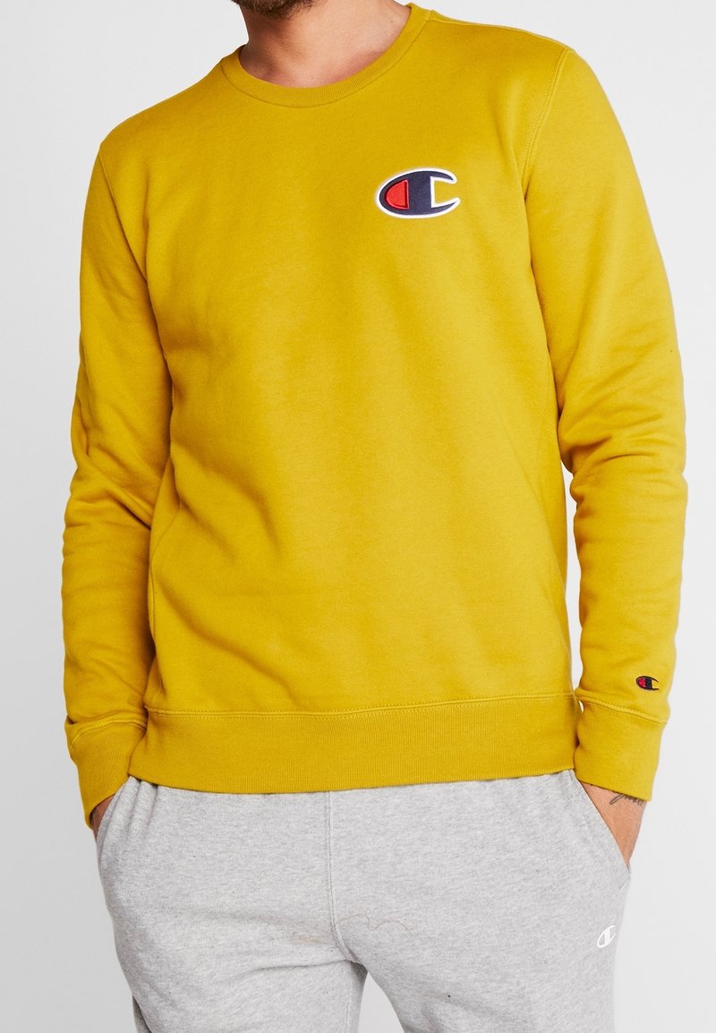 Sudadera amarilla de mezcla de algodón, con puños y dobladillo acanalados, y un pequeño logo bordado en el pecho. El modelo lleva pantalones grises.