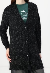 Svart stickad cardigan med v-ringning, chunky tekstur, långa ärmar och fem knappar framtill. Bärs över en grön skjorta.