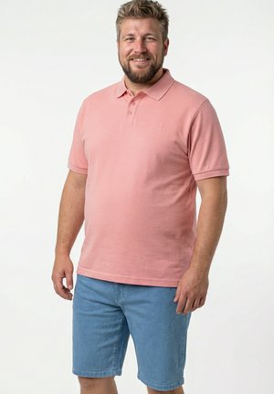 Hombre sonriente con pelo corto rubio y barba, vestido con una camiseta polo rosa y pantalones cortos de mezclilla azul claro, de pie sobre un fondo blanco.