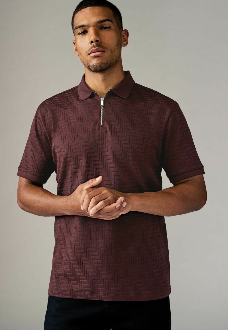 Polo burdeos con cuello con cremallera, que presenta un patrón texturizado de espiga y mangas cortas. El modelo está de pie con las manos entrelazadas.