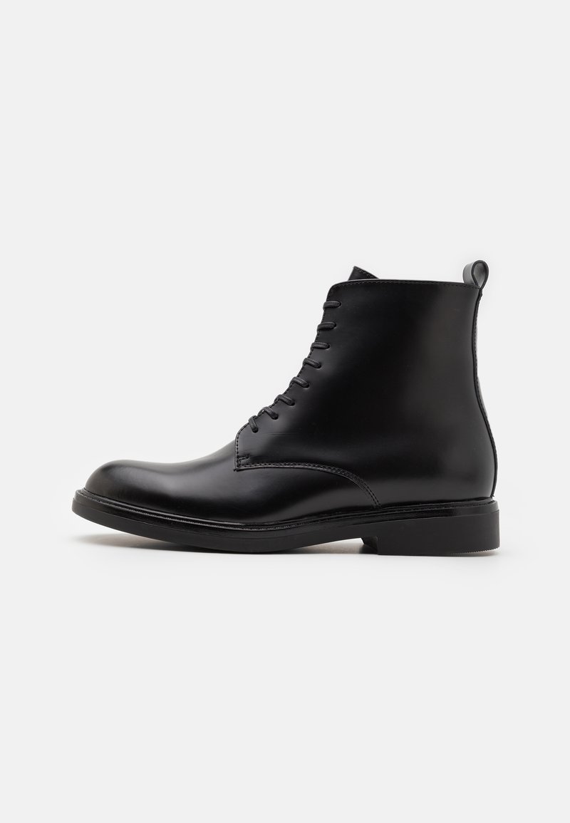 Bottine en cuir noir à bout rond, avec un lacet, texture lisse et un talon solide bas. Dispose d'une languette à l'arrière.