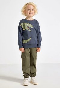 Navy sweatshirt featuring a green dinosaur graphic; paired with olive cargo pants and white sneakers with a textured sole.