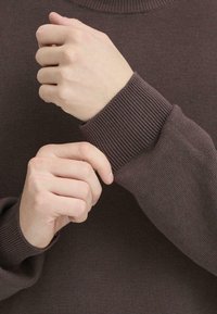 Brown knitted sweater with ribbed cuffs; hands adjusting the cuff. Material appears soft, with a textured pattern throughout.