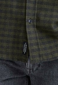 Camisa de franela a cuadros en verde y negro con cierre de botones y una etiqueta de tela en la parte inferior izquierda, combinada con pantalones de mezclilla gris oscuro.