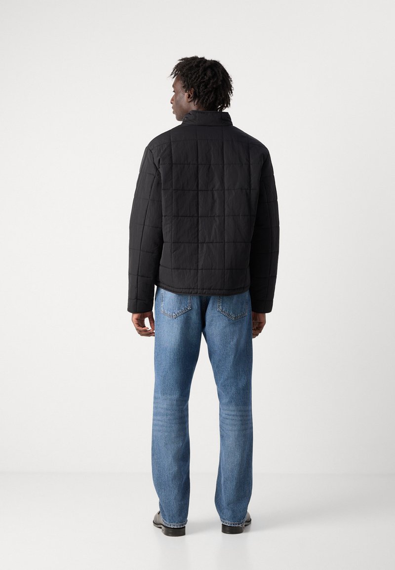 Veste matelassée noire avec un col haut et un motif carré, associée à un jean droit bleu clair et des chaussures noires.