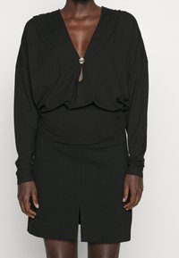 Black long-sleeve top features a deep V-neck, twisted front design, and a decorative button. Paired with a fitted black mini skirt with a slit.