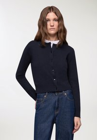 Cardigan navy con bottoni, realizzato in un tessuto morbido, con scollo rotondo e maniche lunghe, abbinato a jeans dritti blu scuro.