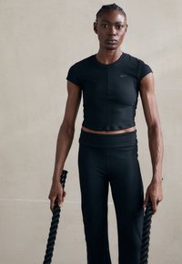 Črn športni crop top in hlače s visokim pasom, obe prilegajoči. Model drži zvite črne vrvi. Gladka tekstura, minimalističen dizajn.