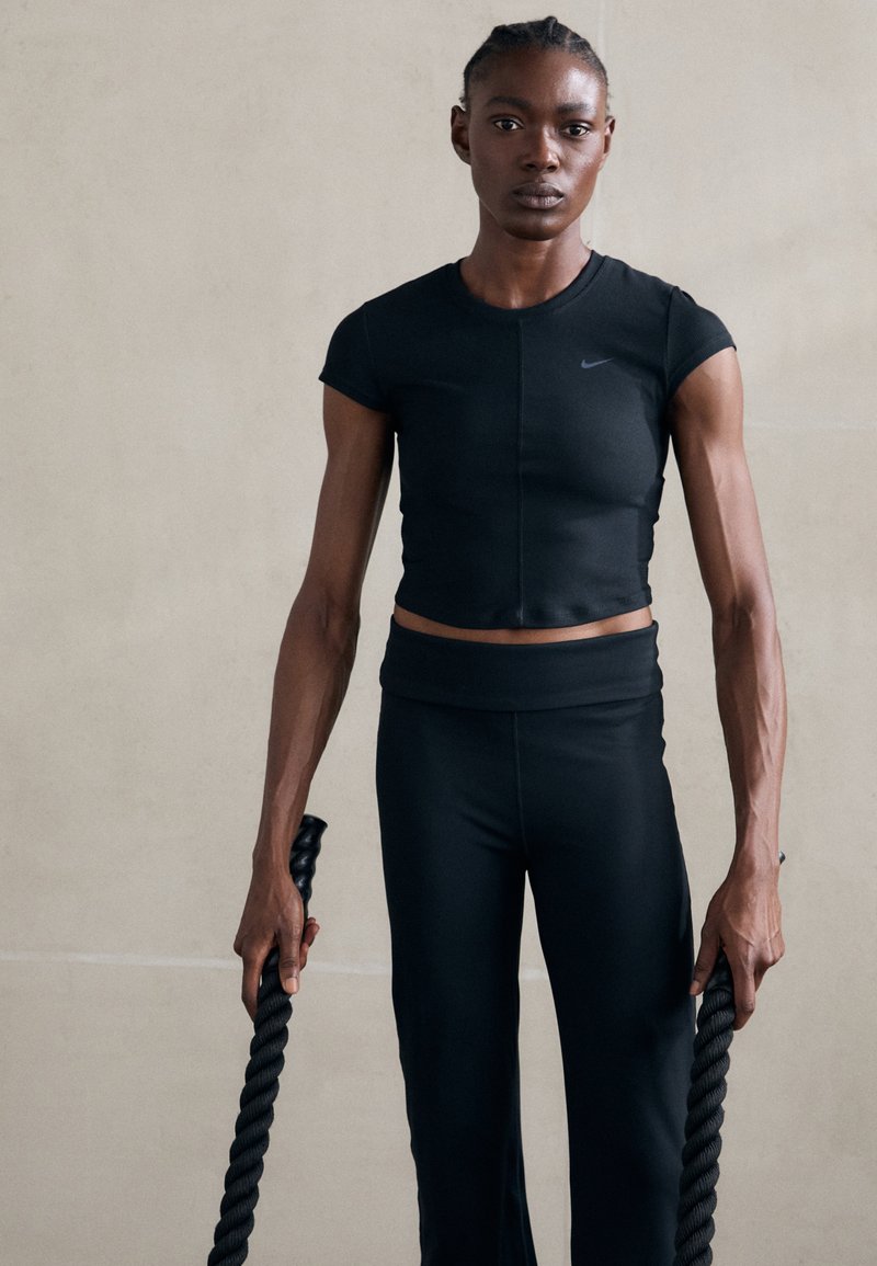Črn športni crop top in hlače s visokim pasom, obe prilegajoči. Model drži zvite črne vrvi. Gladka tekstura, minimalističen dizajn.