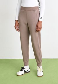 Jogging léger marron clair en tissu doux, avec une taille large, des jambes fuselées et des poches latérales, associé à des baskets blanches et noires.