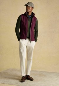 Vesta z burgundskej fleece na zelenej mikine, biele nohavice, hnedé mokasíny a károvaná čiapka. Textúrovaná látka a vrstvený dizajn.