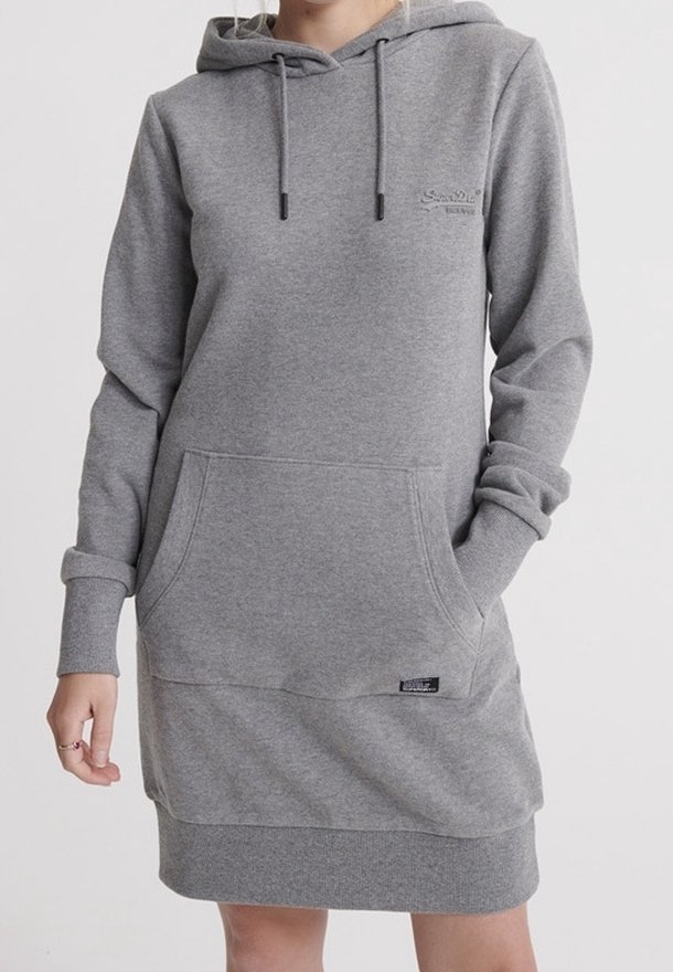 Personne portant une longue robe sweat à capuche grise avec une poche kangourou devant et des poignets côtelés, debout avec une main partiellement dans la poche.