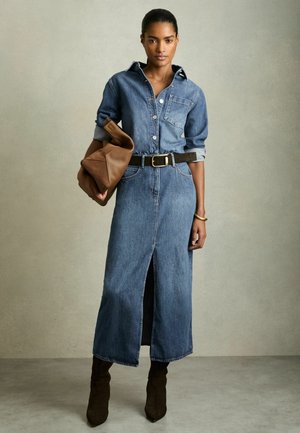 Kvinde iført en lang denimkjole med frontslids, sort bælte, brune støvler, holder en stor brun læder clutchtaske.