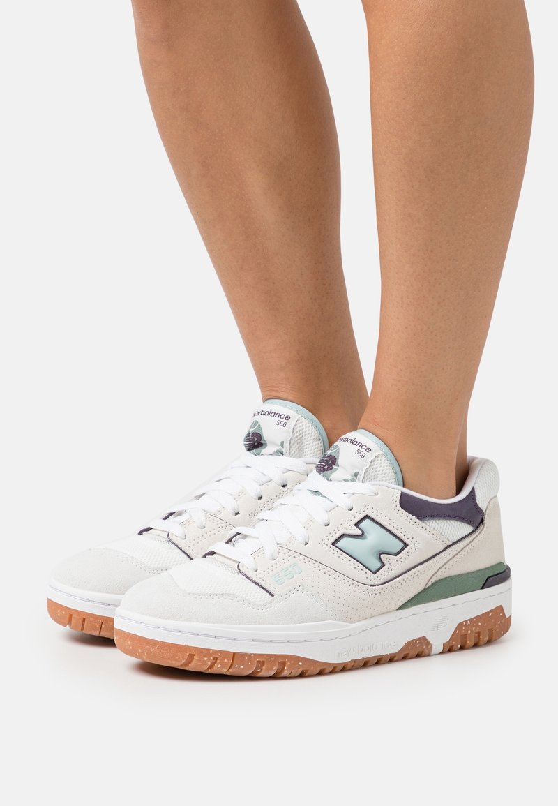 Jambes portant des baskets New Balance 550 blanches et bleu clair avec semelles en caoutchouc, debout sur un fond blanc uni.