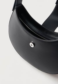 Μαύρη δερμάτινη τσάντα ώμου με καμπυλωτό σχήμα, ορατό φερμουάρ και στρογγυλό ασημί μαγνητικό κούμπωμα μπροστά.