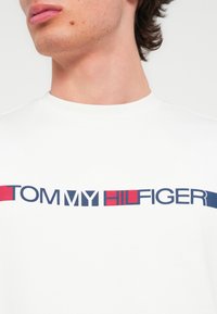 Λευκό βαμβακερό t-shirt με έντονα μπλε και κόκκινα γράμματα που γράφουν "TOMMY HILFIGER." Στρογγυλή λαιμόκοψη με λεία υφή.