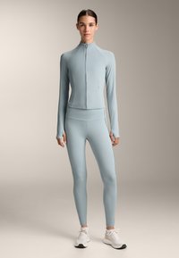 Svijetloplava atletska zip-up jakna s rebrastom teksturom, uparen s uskim tajicama koje imaju bočne šavove i naglaske. Bijele tenisice.