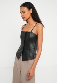 Kortsukkeliste juuste ja musta nahast nööbitud topi ning beežide pükstega naine, seismas tasa valge taustaga.