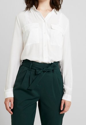 Femme portant une chemise blanche à boutons avec poches poitrine et un pantalon vert foncé taille haute, noué avec une ceinture à nœud.
