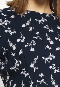 Marinblus med spridda små vita blommönster och rund halsringning, som visar delar av en kvinnas hals och axel.